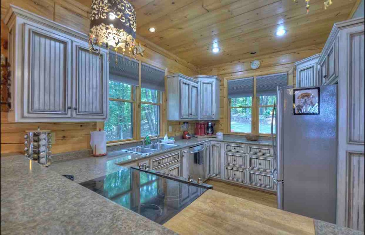 An upscale kitchen features custom cabinetry with a light finish, spacious counter areas, and modern appliances. Natural light fills the room through multiple windows, enhancing the wood-paneled walls. A decorative chandelier adds a touch of elegance, complementing the warm tones of the space.