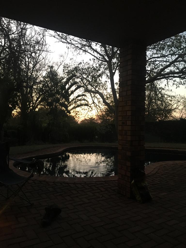 The image captures a serene evening scene by the pool, with reflections of trees and a sunset illuminating the water's surface. Surrounding greenery is visible, along with a brick pillar supporting the roof, creating a sheltered outdoor area.