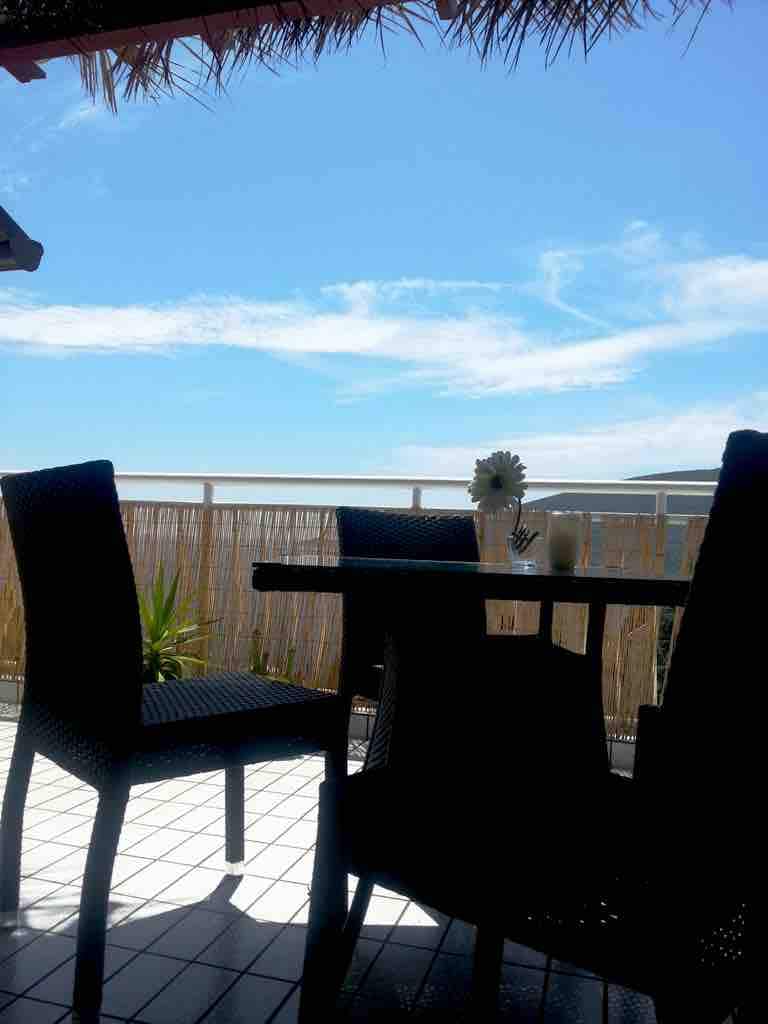A table and four chairs are positioned on a balcony with a clear view of the blue sky. Sunlight frames the scene, emphasizing the openness. Soft greenery and a straw roof are visible in the background, creating a relaxed outdoor space.