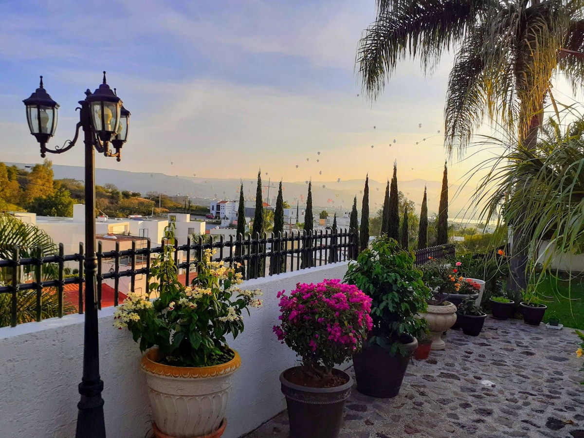 A well-maintained outdoor area is framed by a decorative iron fence. Potted plants in vibrant colors, including bougainvillea and various flowers, are positioned along the walkway made of smooth stones. Palm trees are visible, with a scenic landscape of hills in the background.