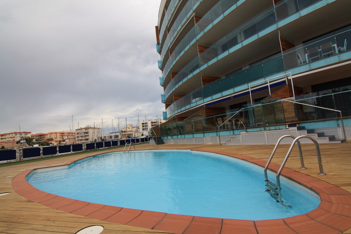 A community pool is situated adjacent to a modern apartment building, featuring a circular design with a wooden deck. Steps lead into the water, which appears calm. Surrounding the pool, views of neighboring buildings and marina can be seen, offering a serene environment.