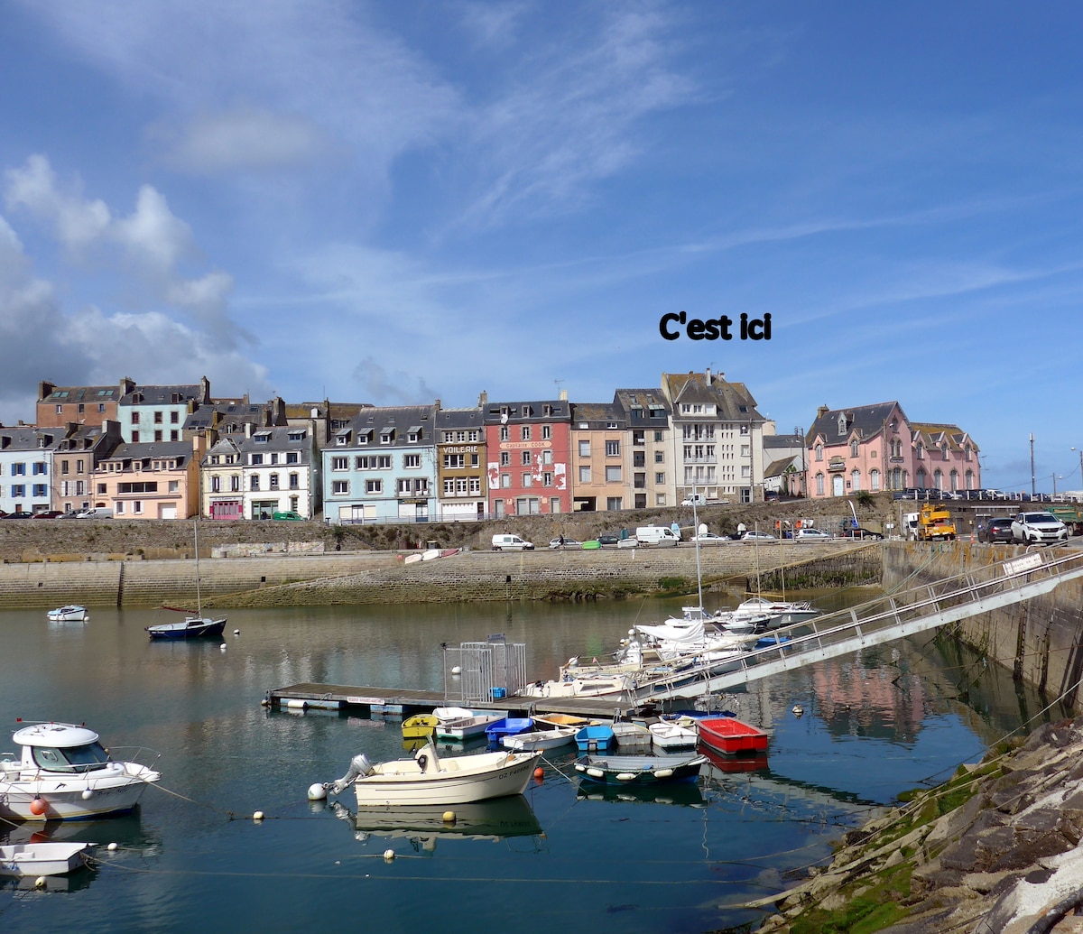 The image displays a charming harbor scene, featuring several small boats moored in calm waters. Colorful buildings line the waterfront, showcasing a variety of hues including pastel pinks and blues. The sky is clear with a few scattered clouds, providing a bright backdrop.