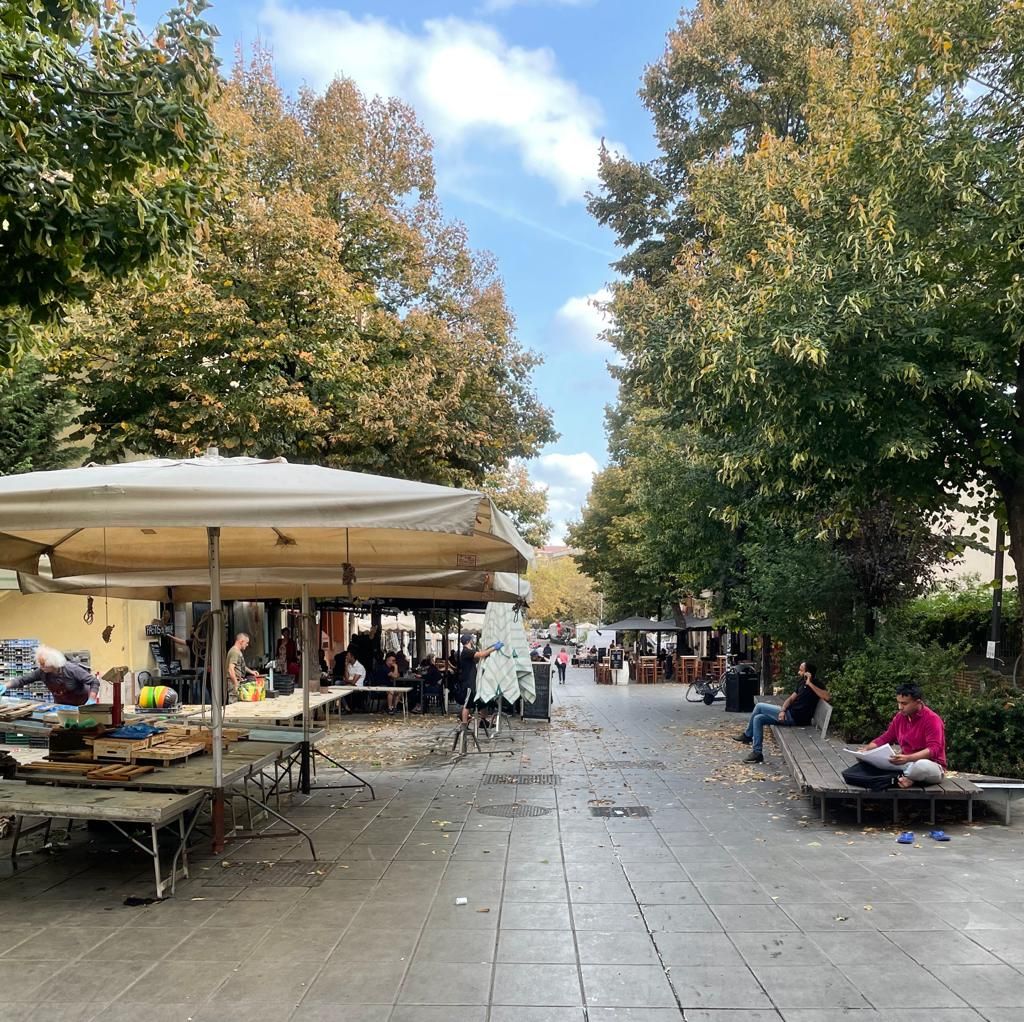 A pedestrian area filled with market stalls is lined with trees displaying autumn foliage. Umbrellas provide shade over tables displaying various items. Benches are located along the sides, inviting relaxation amidst the activity. The sky is clear with a few scattered clouds.