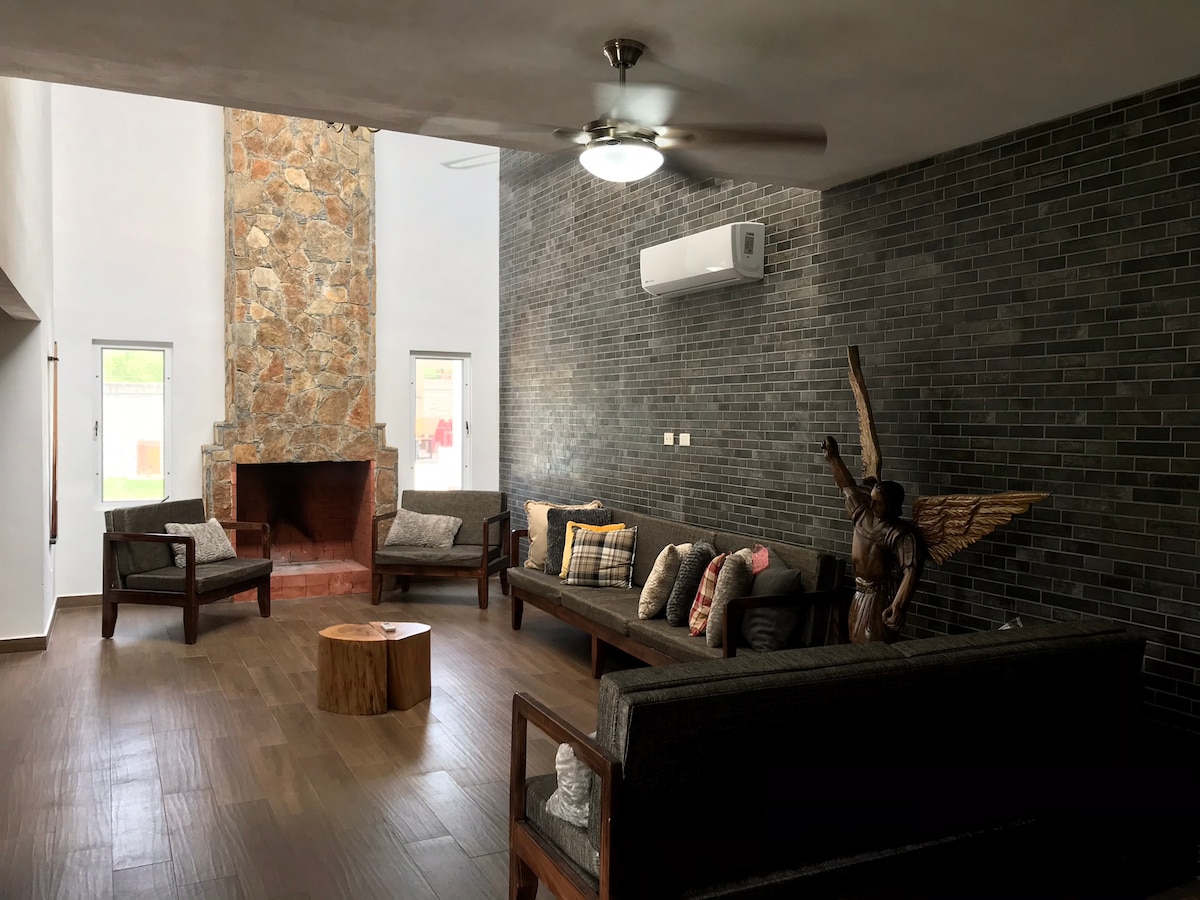 A spacious living area features a striking stone fireplace as a focal point. Comfortable seating is arranged around a wooden accent table, with decorative pillows enhancing the ambiance. Large windows invite natural light, complemented by modern ceiling fans and an air conditioning unit.