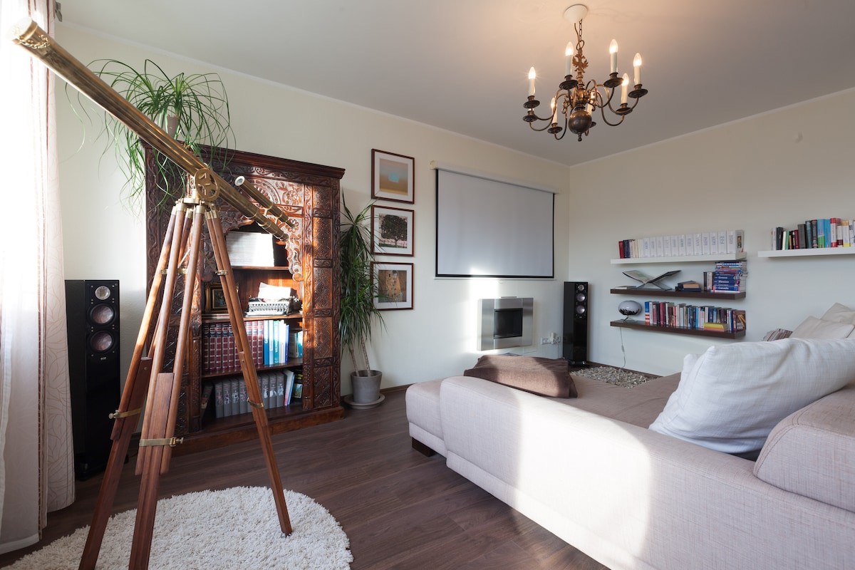 A spacious living area is highlighted by a classic telescope on a wooden stand. A stylish chandelier hangs above, while a cozy sectional sofa is positioned near a rug. Shelves with books and decorative items are visible, along with a wall-mounted screen and a sleek fireplace.