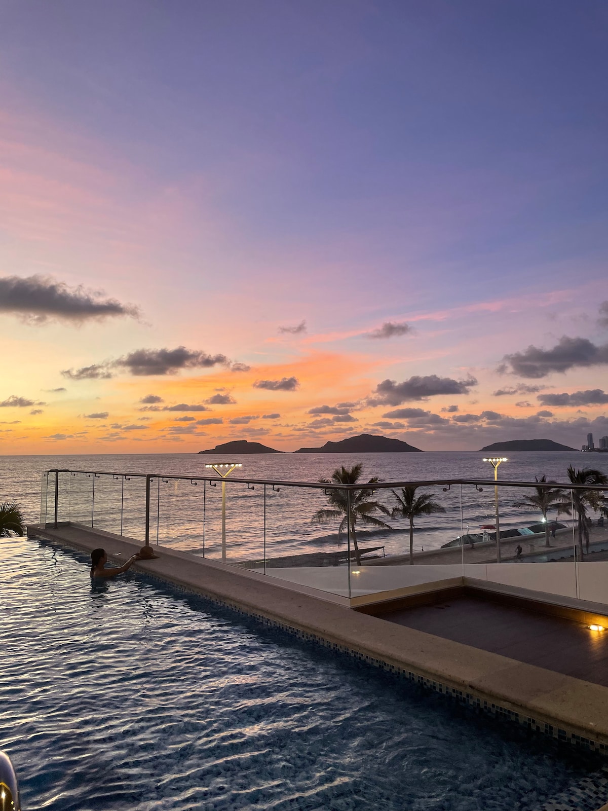 An infinity pool merges seamlessly with the ocean, reflecting the vibrant colors of a sunset. Silhouettes of palm trees are visible along the shoreline, while distant islands enhance the serene coastal view. Soft lighting along the pool edge adds an inviting ambiance.
