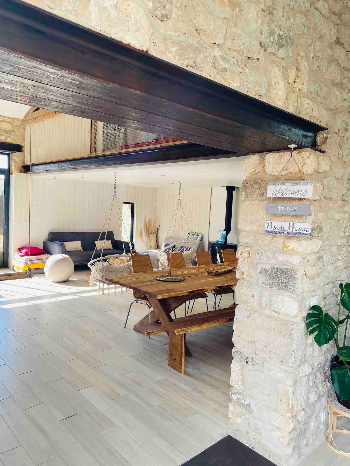 A spacious living area is highlighted by a rustic stone wall and wooden beams. A large dining table accommodates multiple guests, while comfortable seating options, including sofas and a hanging chair, invite relaxation. Natural light floods the space, enhancing the inviting environment.