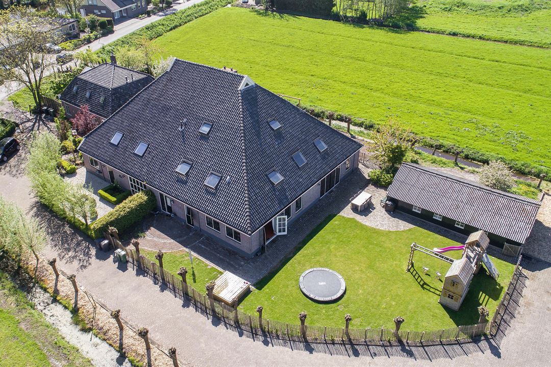 An aerial view of a spacious farm property is captured, showcasing a large building with a sloped roof. Surrounding green pastures and a lawn featuring a small playground and trampoline are visible. The property is well-maintained, with hedges lining the driveway.