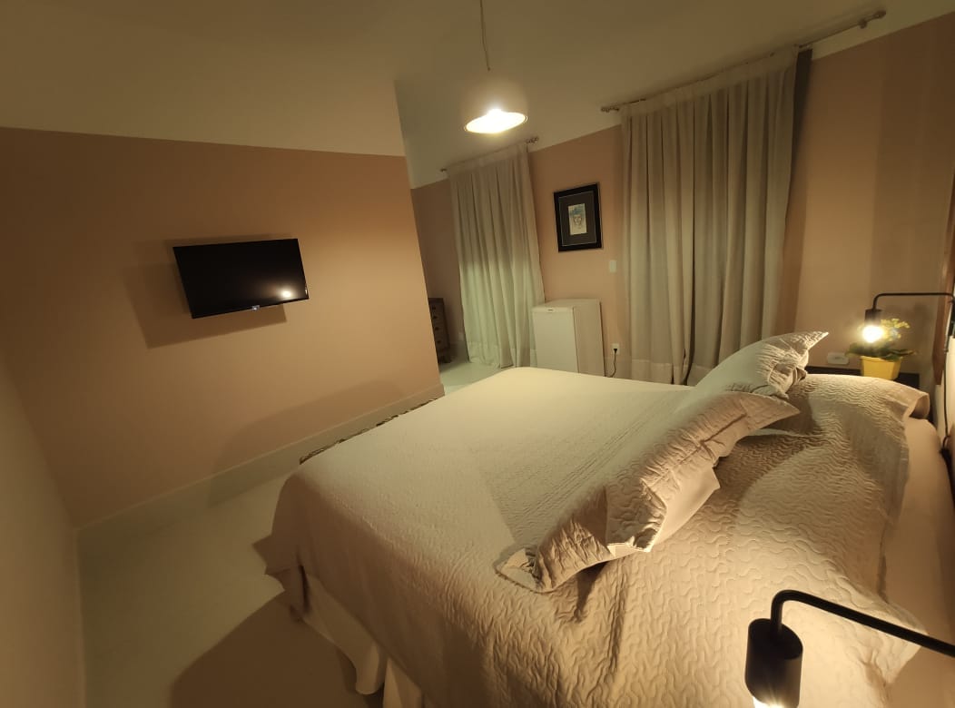 A softly lit bedroom is displayed, featuring a spacious bed with light-colored bedding. A flat-screen television is mounted on the wall opposite the bed. Elegant curtains frame the windows, and a small dresser is positioned near the room's entrance.