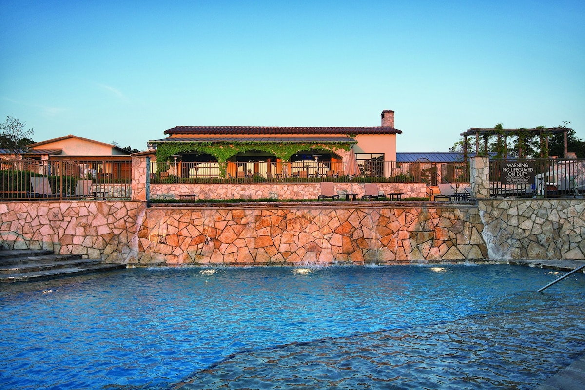 A multi-level outdoor area offers a stone wall surrounding a sparkling pool, with sun loungers nearby. A covered terrace is visible in the background, featuring a wooden structure adorned with greenery. The setting is illuminated by the warm tones of sunset.