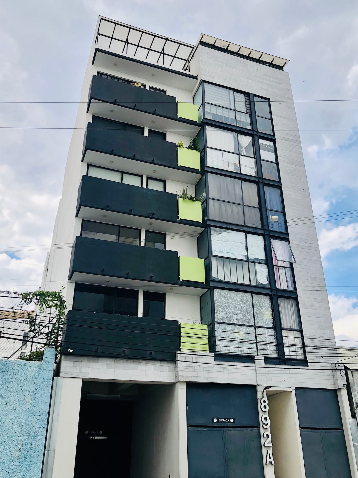 A modern building features five stories, with a combination of black and green balconies. Large glass windows allow natural light to enter the structure. The entrance is visible at the bottom, showcasing a sleek, minimalistic design that complements the overall aesthetic.