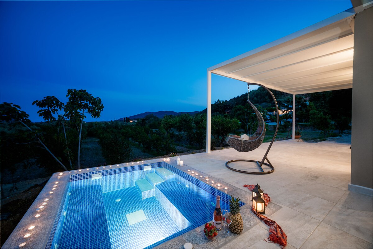 An outdoor patio features a private jacuzzi illuminated by soft lighting, surrounded by serene landscaping. A comfortable hanging chair is positioned nearby, creating a relaxing space. The twilight sky casts a calming ambiance over the area, enhancing the peaceful surroundings.