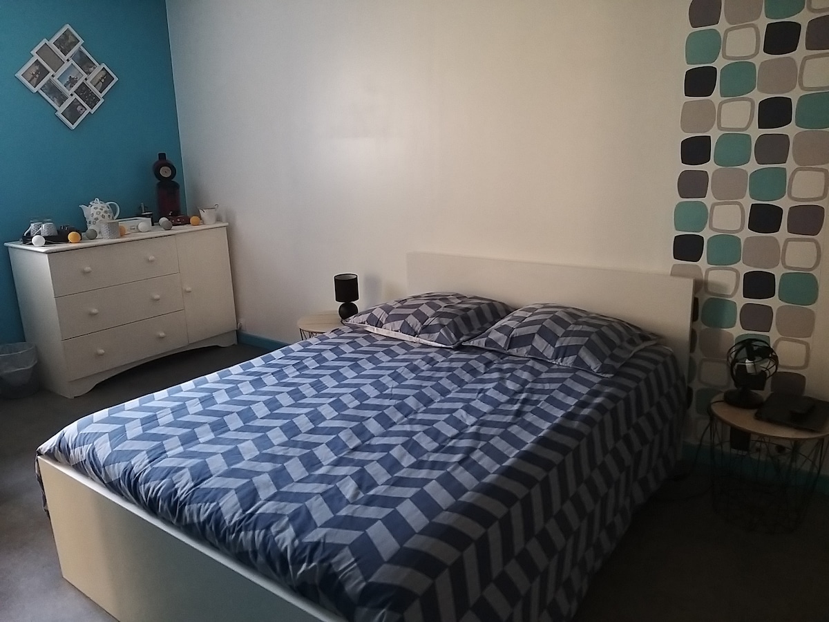 A private bedroom features a spacious bed covered with a blue and white patterned blanket. A light-colored dresser stands against the wall, and a small bedside table is positioned beside the bed. The walls are painted in soft blue tones with geometric accents.