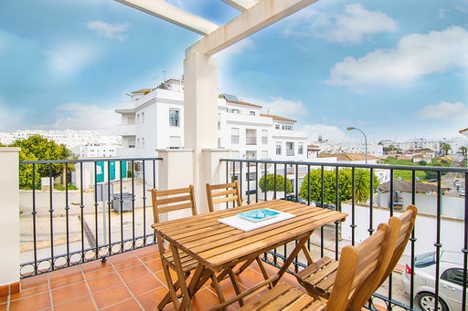 An outdoor terrace is featured, furnished with a wooden table and four matching chairs. Light-colored walls and a quaint railing enhance the inviting atmosphere. Surrounding trees and buildings are visible in the background, providing a glimpse of the local neighborhood.