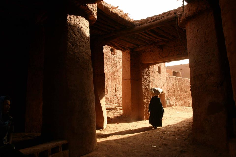 A sunlit pathway is viewed through a rustic archway, revealing textured earthen walls and supporting pillars. The scene captures a solitary figure walking gracefully, with traditional attire and a sunshade. Natural light creates soft shadows on the ground, enhancing the serene desert ambiance.