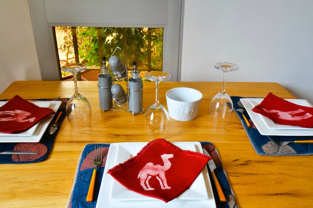 A dining table is set with white plates, red napkins featuring a camel design, and elegant wine glasses. A minimalist centerpiece with condiments is positioned between the place settings, while natural light filters through a window showcasing greenery outside.