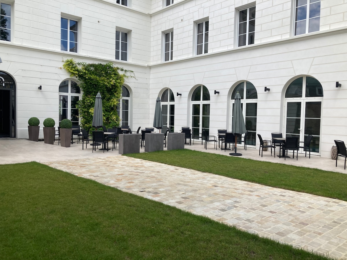 An outdoor seating area is presented, featuring several dark metal tables and chairs arranged on a stone pathway. Two large umbrellas provide shade, while lush greenery complements the light-colored building façade. The area invites relaxation in a serene garden setting.