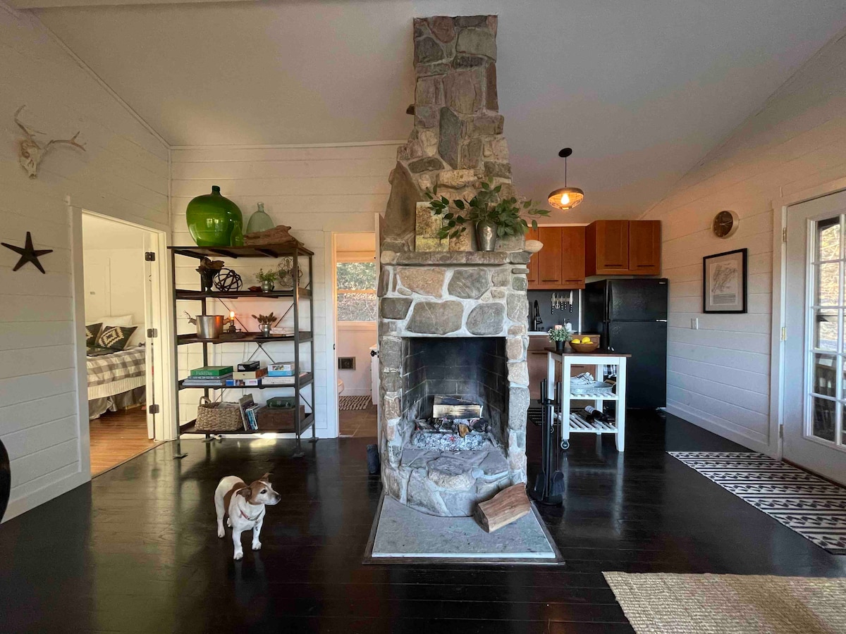 A stone fireplace is the focal point of the cozy main room, surrounded by wood flooring and natural light. Open shelving displays decorative items. A small kitchen features wood cabinetry, while a dining area is visible in the background, enhancing the inviting layout.