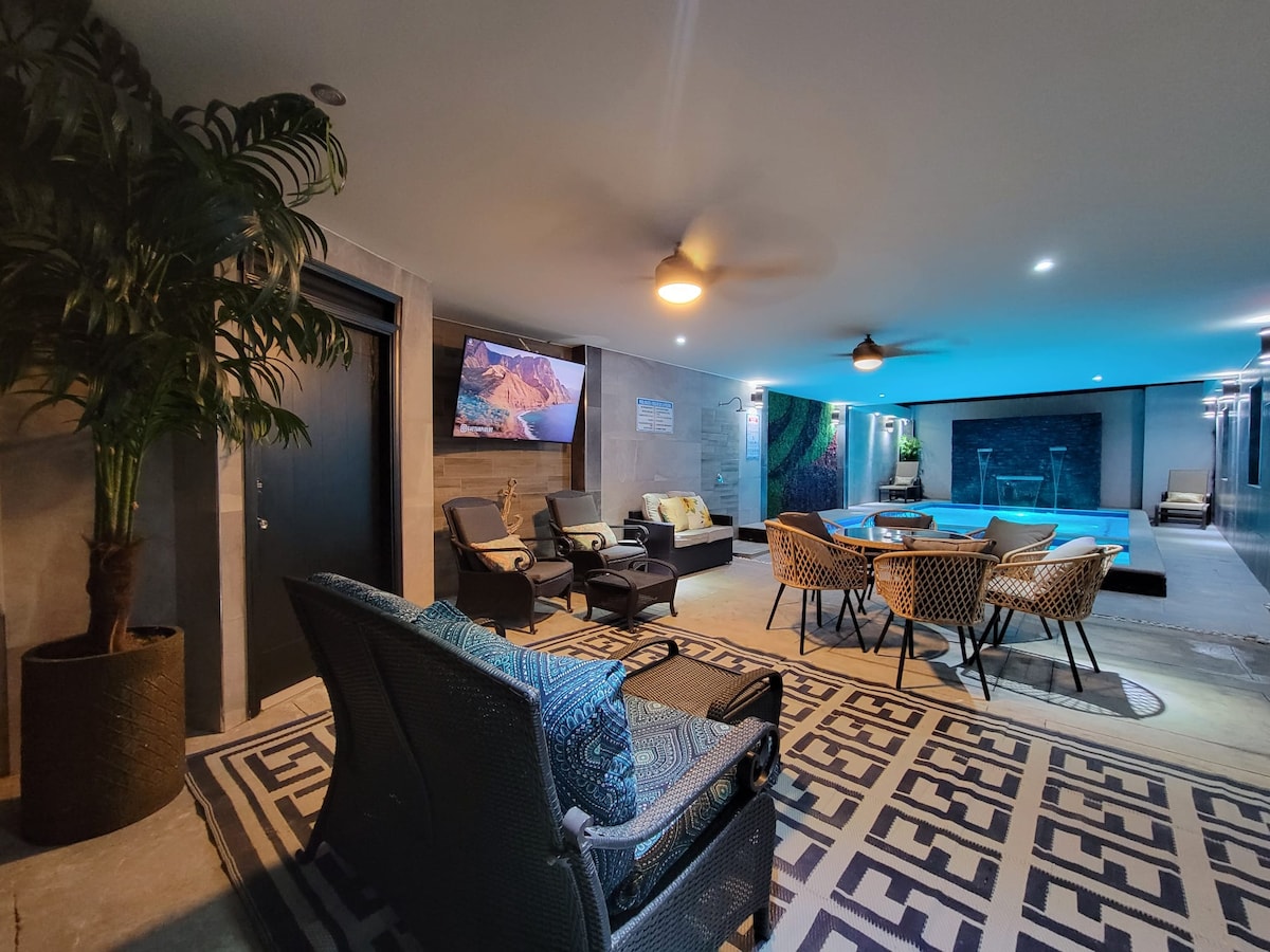 An inviting indoor space features a comfortable seating area with dark wicker chairs and light-colored cushions. A patterned rug spans the floor, while a wall-mounted screen and ceiling fans provide modern amenities. An illuminated pool area can be seen in the background, enhancing the ambiance.