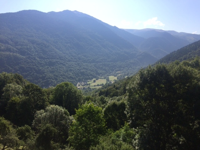 A wide view of the lush valley is captured, surrounded by rolling mountains. Verdant trees and open fields are visible beneath the bright blue sky, with soft clouds scattered above. The landscape showcases the natural beauty of the area, inviting exploration and relaxation.