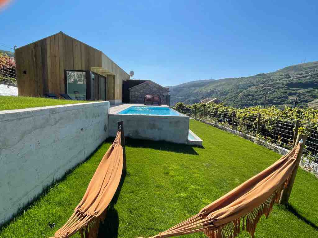 A spacious lawn is set in front of a contemporary house, featuring a clear blue pool surrounded by wooden deck chairs. Two hammocks are positioned beneath the sunlight, with a backdrop of rolling green hills and vineyard rows, creating an inviting outdoor retreat.