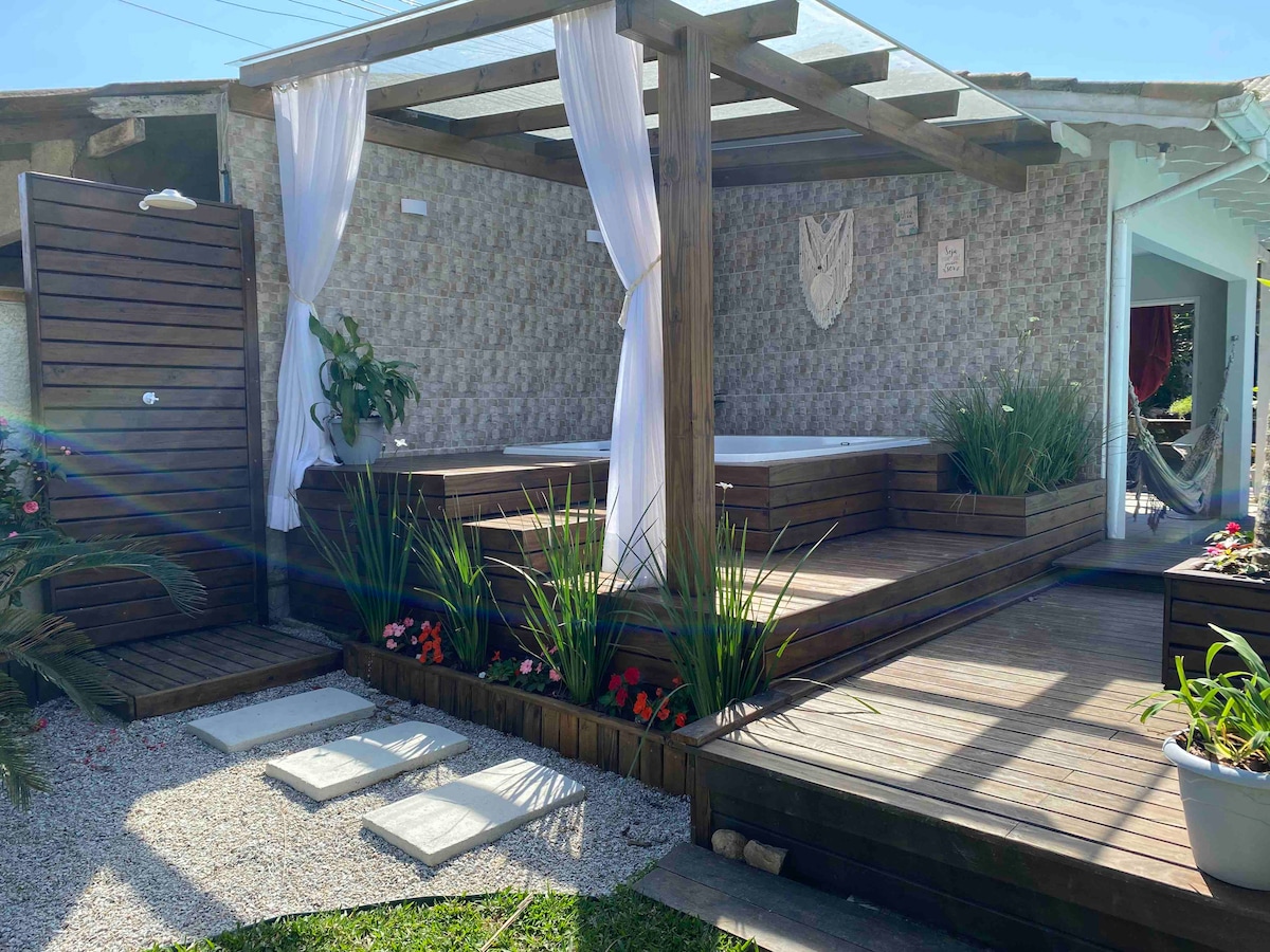 An outdoor space is presented, featuring a wooden deck surrounded by plants and flowers. A jacuzzi is nestled within the deck area, covered by a transparent roof. White curtains drape elegantly, enhancing the serene atmosphere. Pathway stones lead to the jacuzzi area, promoting accessibility.