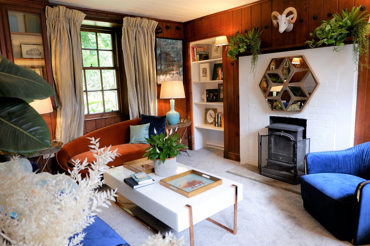 The living room features original oak paneling and a fireplace surrounded by a geometric mirror. Soft furnishings include an orange velvet sofa, a blue accent chair, and a coffee table topped with decorative items. Natural light filters through the window, illuminating a variety of plants.