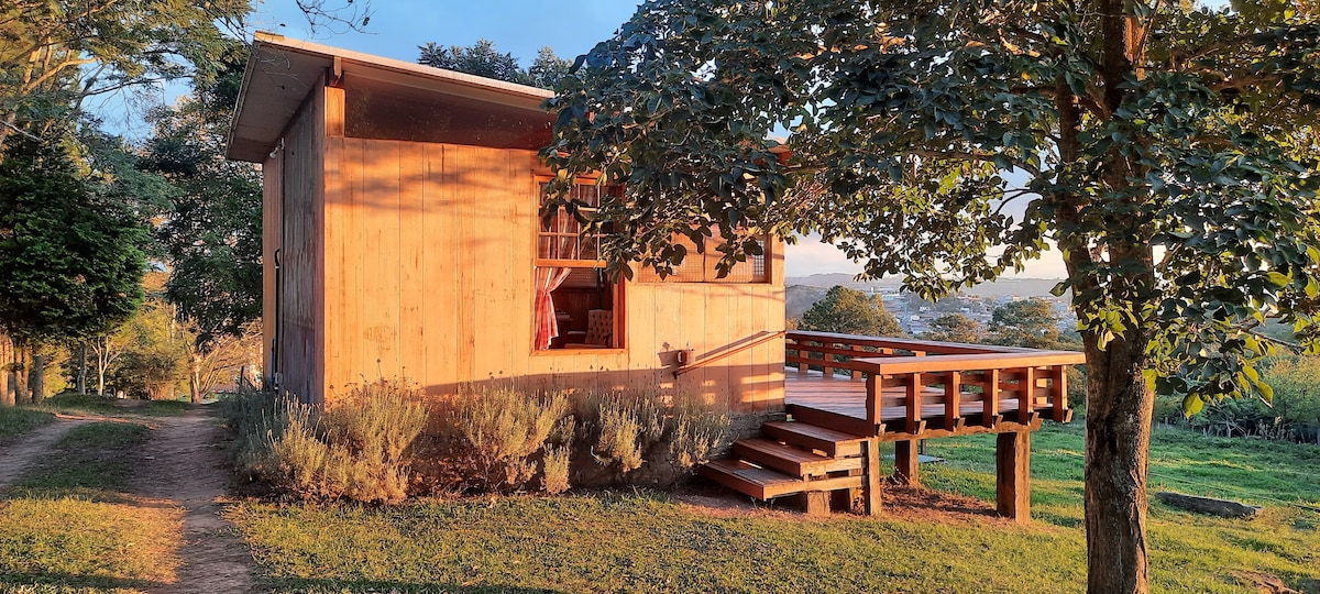 A wooden lodge is set in a lush green landscape, featuring a spacious deck that extends from the entrance. Large windows allow for natural light to fill the interior, while nearby trees provide shade and enhance the connection to nature.
