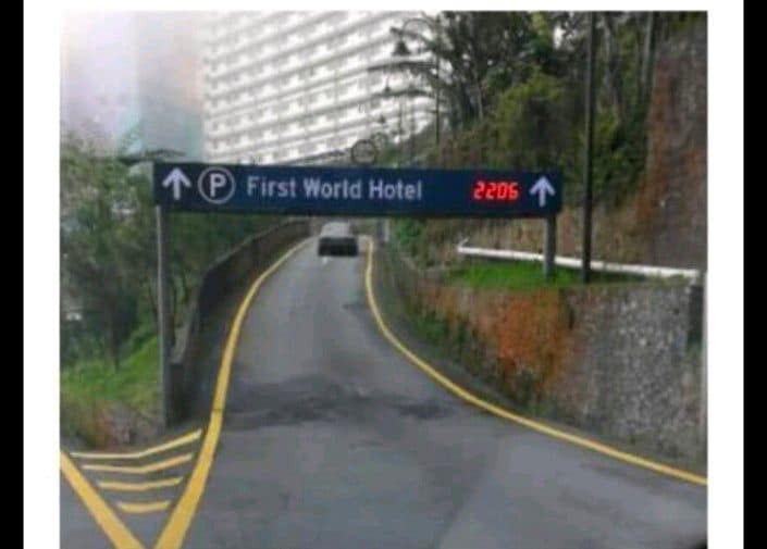 A road sign indicates the direction to the First World Hotel, positioned above a winding pathway. The nearby landscape features greenery and a portion of the hotel's multistory building is visible in the background amidst a foggy environment.