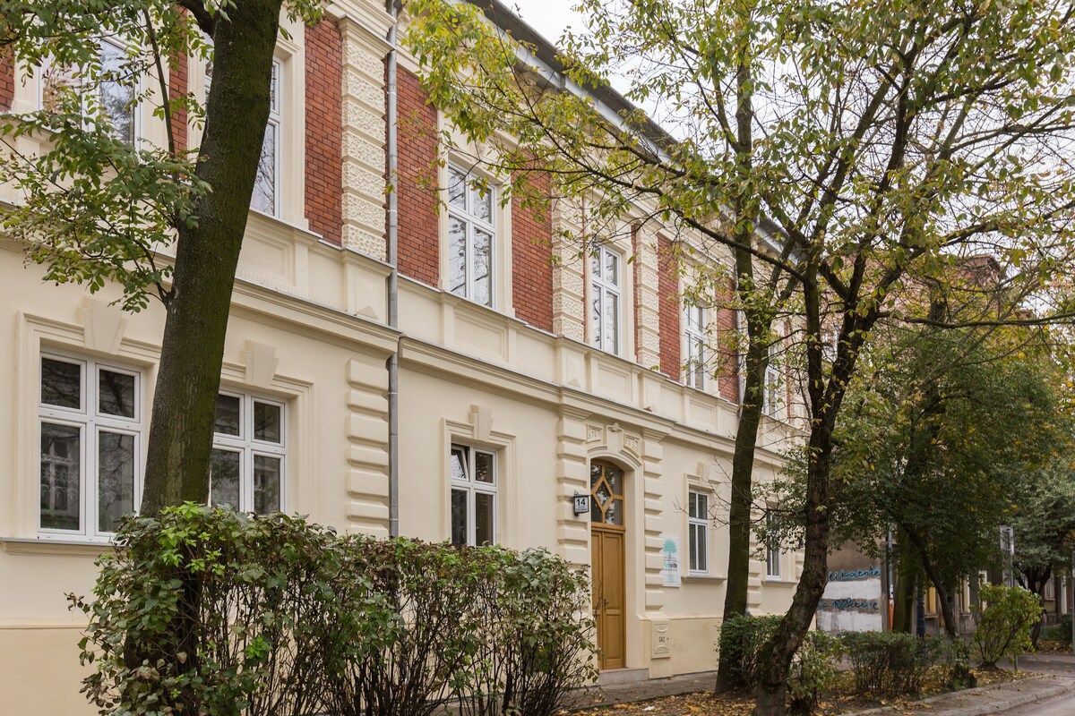 A historic 19th-century townhouse is showcased, featuring a classic exterior with red brick accents and large windows. Trees line the front, adding greenery to the scene, while neat hedges enhance the inviting atmosphere of the building.