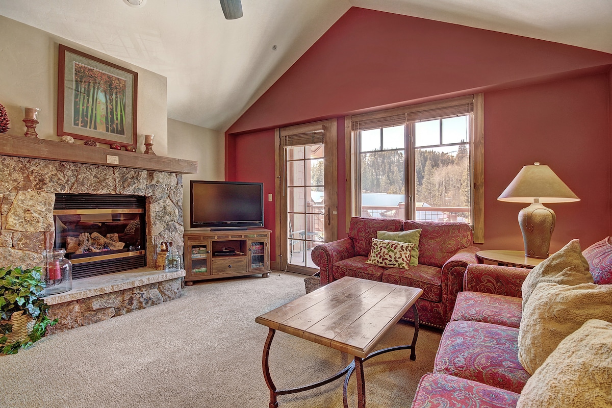 A cozy living area features a stone fireplace with log accents. Two plush sofas provide comfortable seating, while a wooden coffee table sits at the center. Large windows frame the space, allowing natural light to enhance the warm tones of the decor.