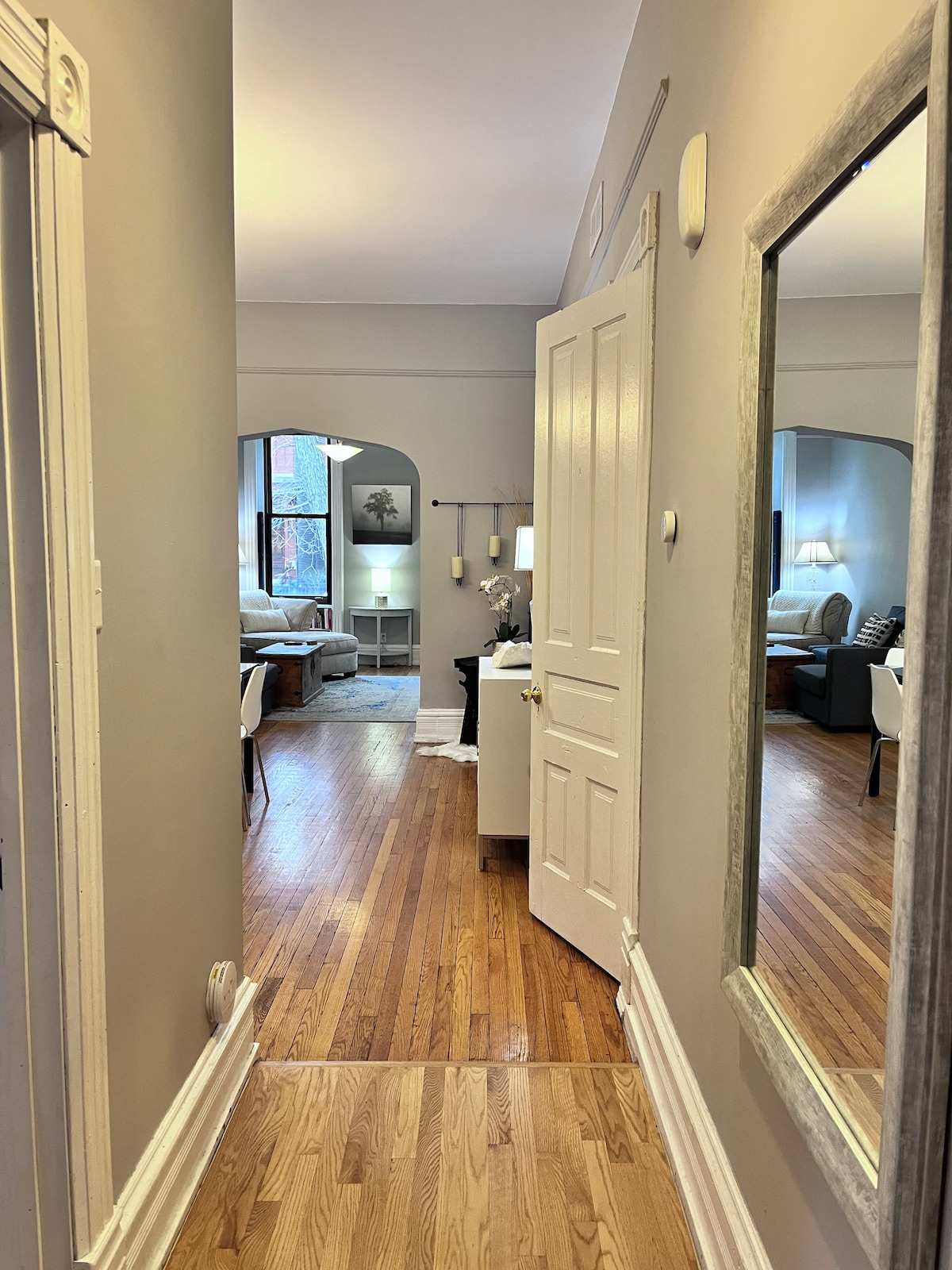 A hallway opens to a living area, featuring smooth hardwood floors and neutral-colored walls. A decorative mirror is mounted near the entrance. Light from a window bathes the space, highlighting various furnishings like seating and side tables within the visible living room.