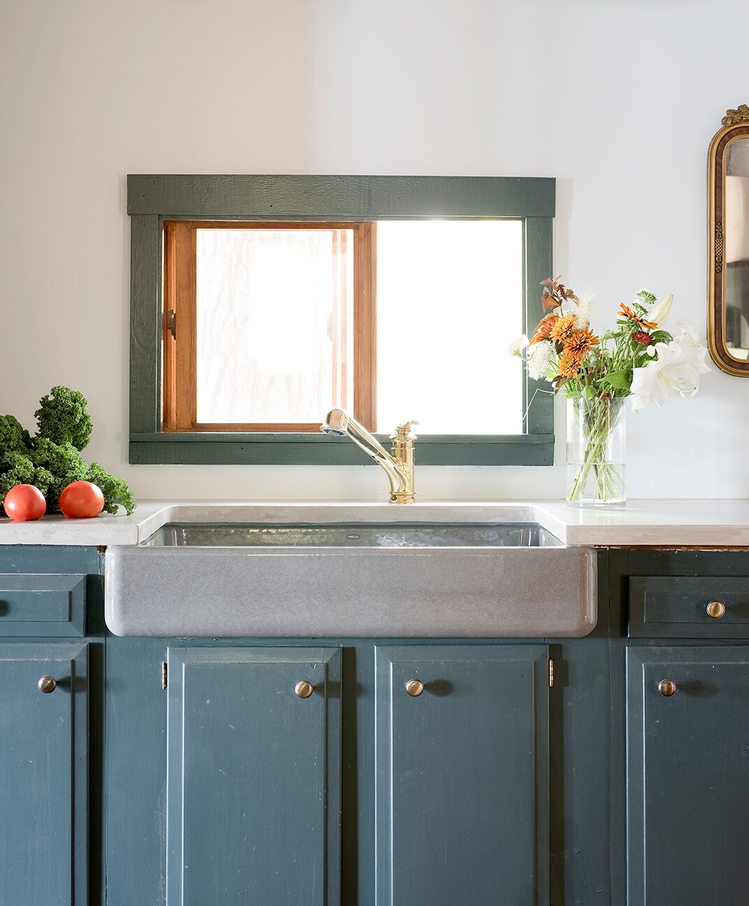 A farmhouse-style sink is positioned beneath a small window, allowing natural light to brighten the space. Fresh vegetables and a vase of flowers add a touch of color to the countertop, which complements the dark, cabinetry beneath. A gold faucet contrasts elegantly with the muted colors.