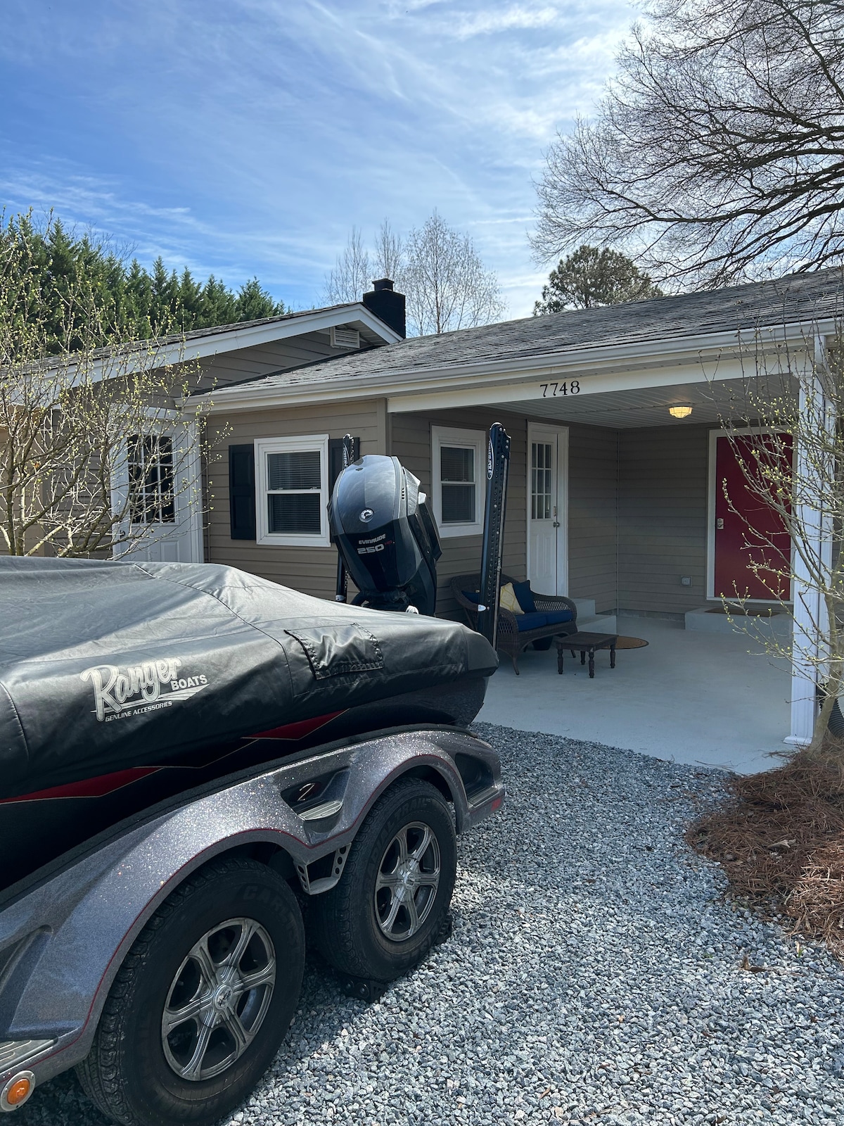 The exterior of the home is showcased, featuring a gravel driveway with a boat attached to a trailer. A covered porch area is visible with seating, complemented by greenery and trees in the background. The home's entrance is framed by light-colored siding and a bold red door.
