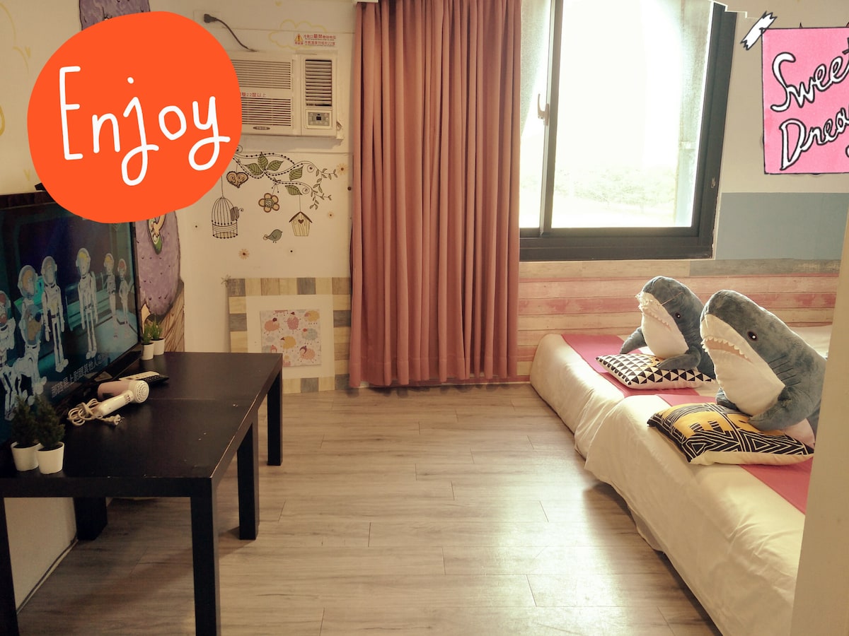 A cozy room features a spacious bed with playful plush shark pillows. A small black table is positioned in front of a television, and a window provides natural light, complemented by light curtains. Decorative art adorns the walls, contributing to a welcoming atmosphere.