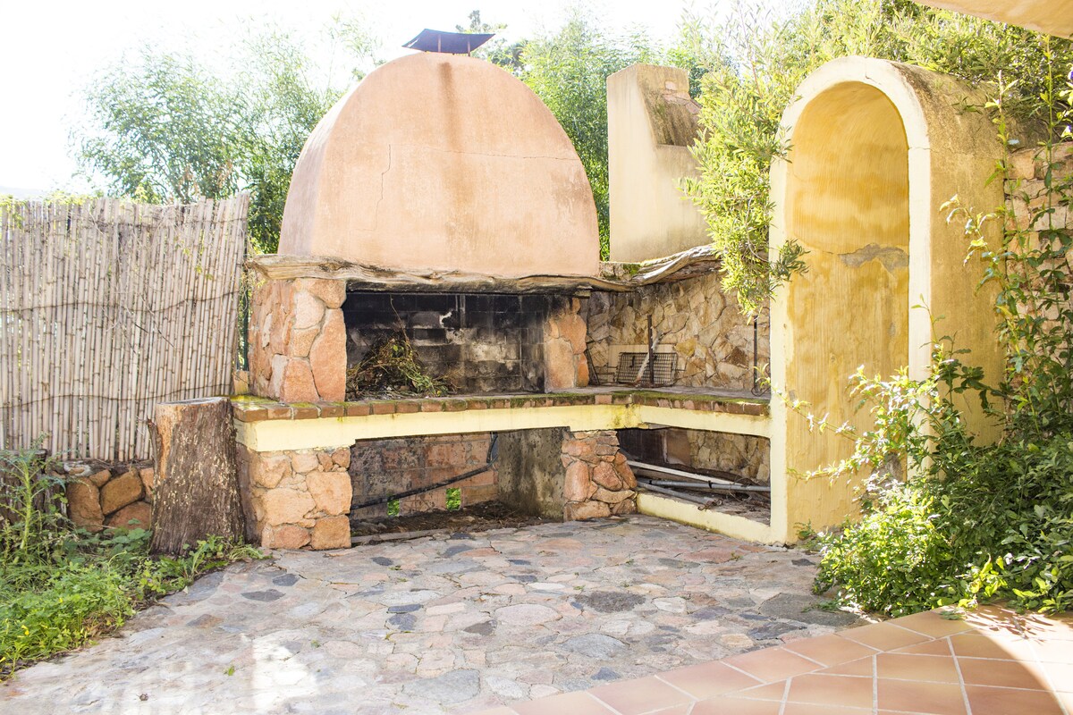 An outdoor barbecue area is featured, constructed with natural stone and a curved brick oven. Surrounded by lush greenery, the space includes a stone patio, providing a rustic atmosphere for dining and social gatherings.