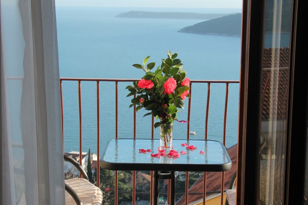 A glass table is set on a balcony, overlooking the blue waters of Boka Bay. A vase with fresh roses is the centerpiece, with petals scattered around it. The scene captures a peaceful outdoor space framed by a railing and light curtains.