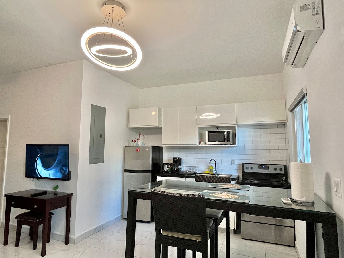 A modern kitchen and dining area is showcased, featuring sleek white cabinetry and stainless steel appliances. A round, contemporary light fixture hangs above the dining table. A television is mounted on the wall, and a window allows natural light to brighten the space.