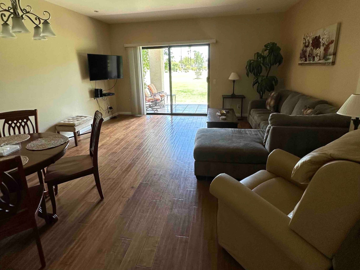 A spacious living area features a large sectional sofa and a flat-screen TV mounted on the wall. A dining table with six chairs is positioned nearby. A glass door leads to the outdoor patio, allowing natural light to brighten the room.