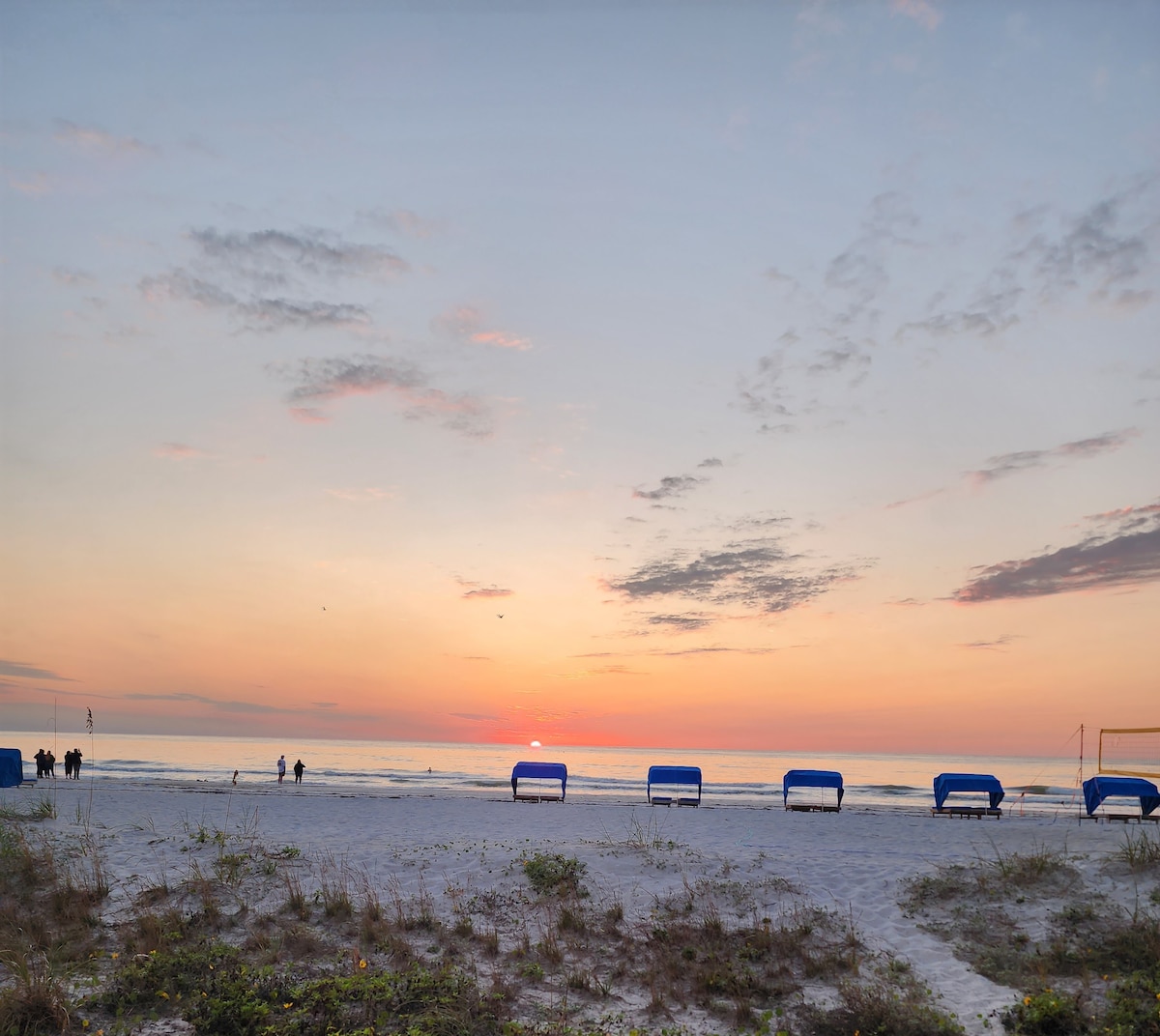 A serene beach scene during sunset is captured, with gentle waves lapping against the shore. Blue beach loungers are positioned in the sand, providing a place to relax. The sky displays soft hues of orange and pink as the sun descends towards the horizon.
