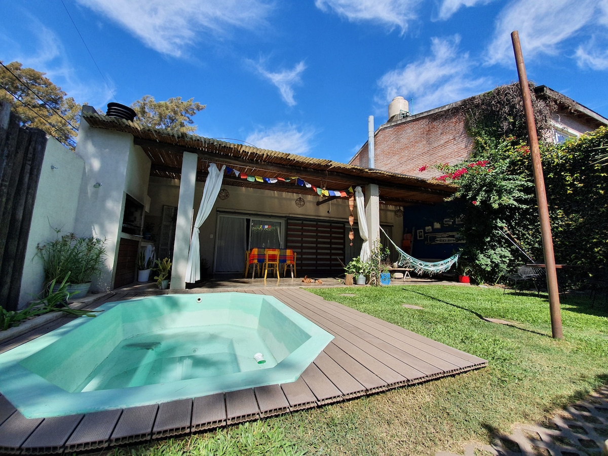 A compact jacuzzi-style pool is surrounded by a wooden deck and lush grass. The outdoor area features a thatched roof and glass doors leading to a spacious interior. Colorful decorations hang above bright chairs, offering a vibrant and relaxing atmosphere.