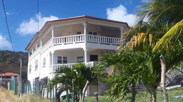 A three-story villa is framed by palm trees and lush greenery, showcasing a white façade with arches and a red-tiled roof. Balconies extend from the second level, providing a view of the surroundings under a clear blue sky.