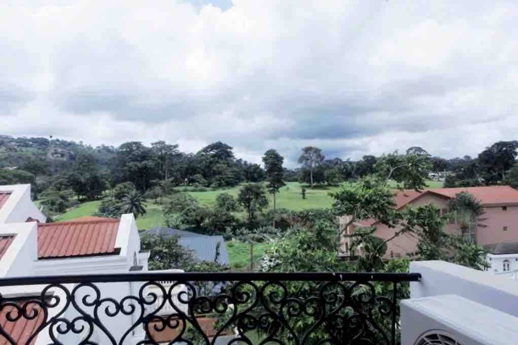 An expansive view from the balcony reveals lush greenery and a golf course beyond. The landscape is dotted with trees and residential buildings, bathed in soft, natural light under a cloudy sky. The decorative balcony railing adds a touch of elegance to the scene.