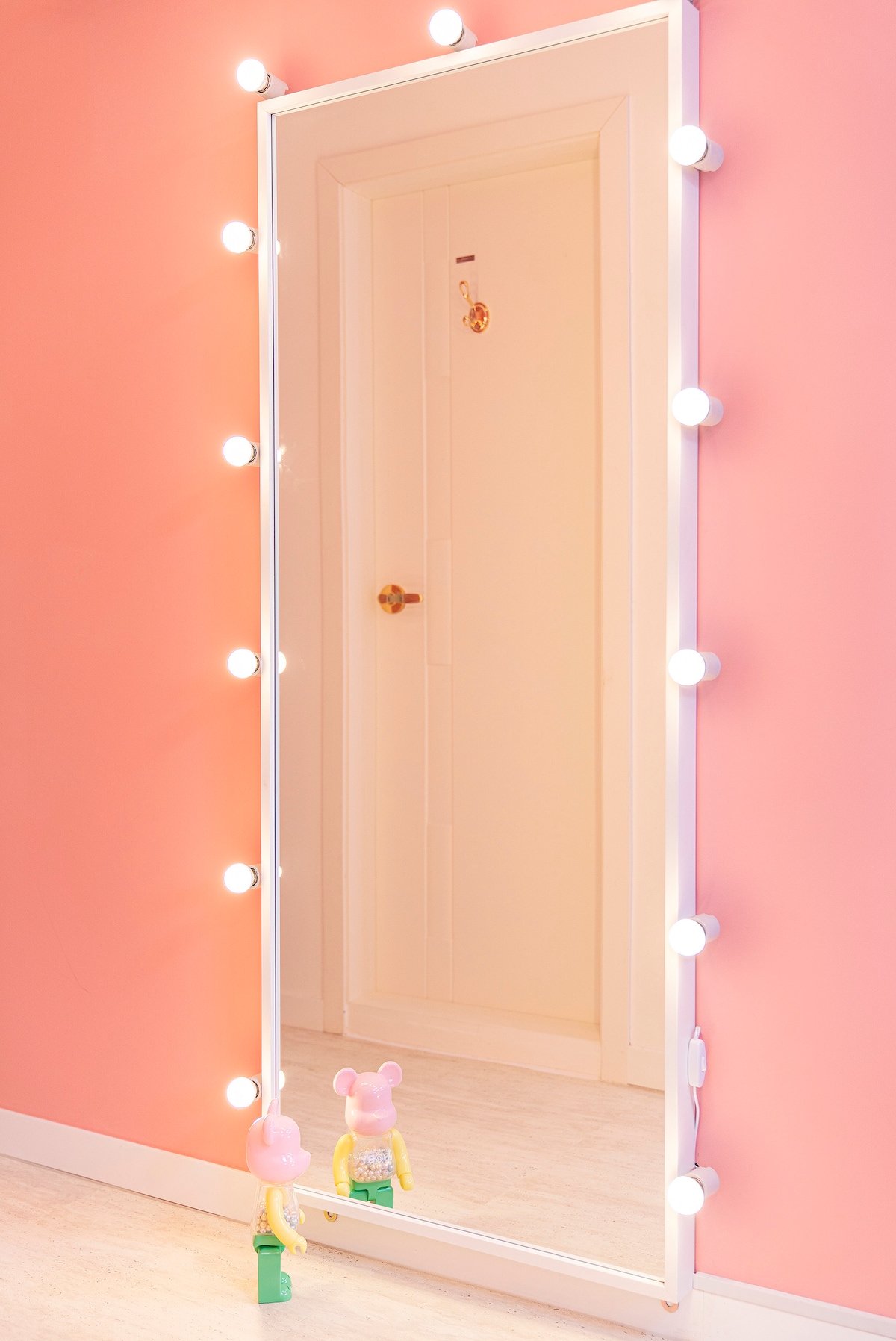 A full-length mirror framed in white stands against a pink wall, illuminated by soft, round bulbs. Colorful toy figures are positioned at the base of the mirror, adding a playful touch. A door can be seen in the background, featuring a golden doorknob.