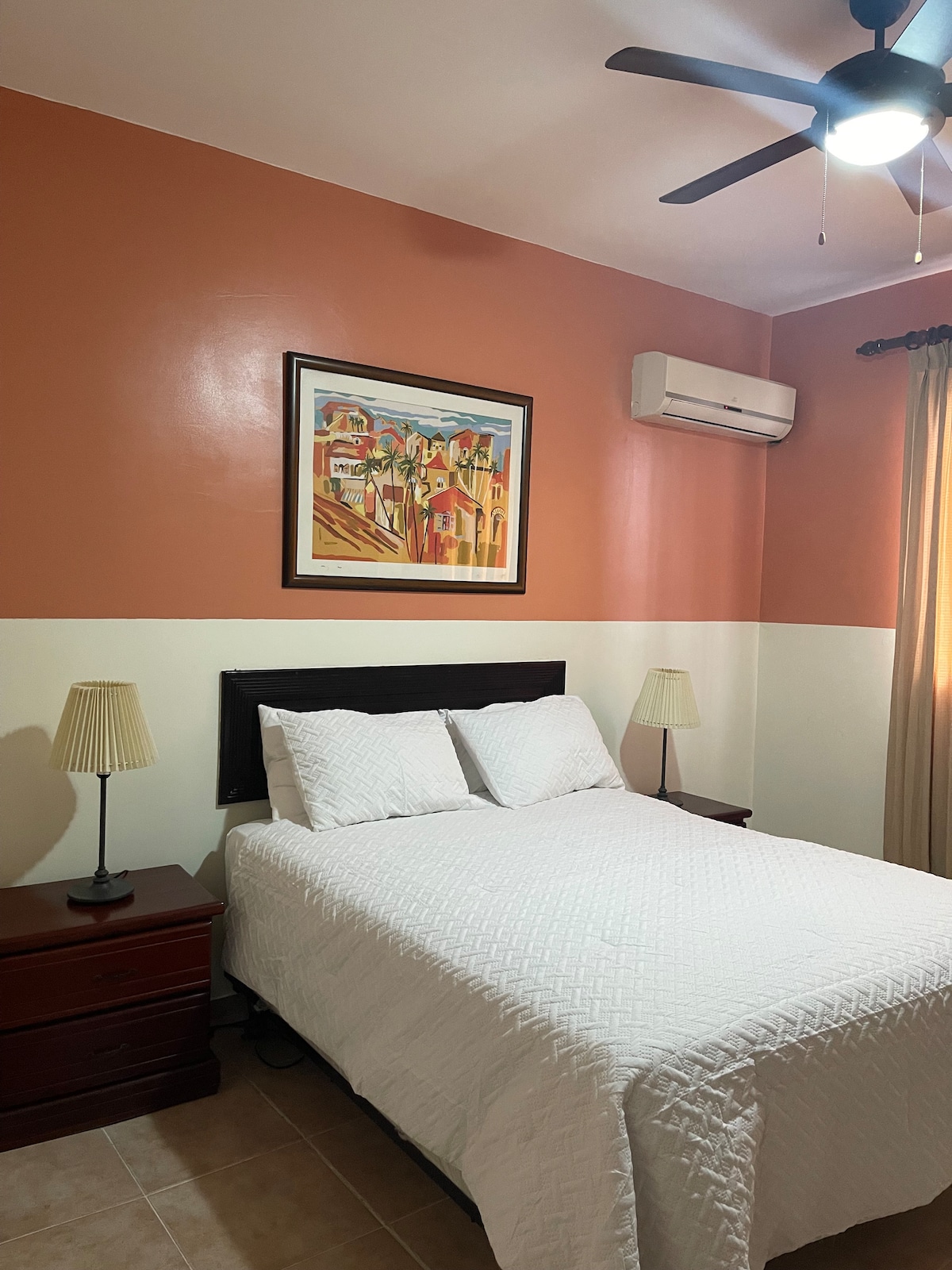 A cozy bedroom features a spacious bed covered with a textured white quilt. Two bedside lamps provide soft light beside the bed, which is positioned against a warm, peach-colored accent wall. An air conditioning unit is mounted above, and a colorful piece of artwork is displayed on the wall.