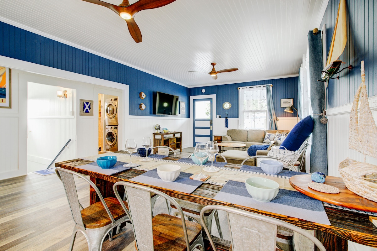 An open living and dining area features a long wooden table set with blue and white dishware. A comfortable seating arrangement is present, including a sofa and a blue chaise lounge. Light-colored walls contrast with nautical-themed decor, while large windows allow natural light to fill the space.