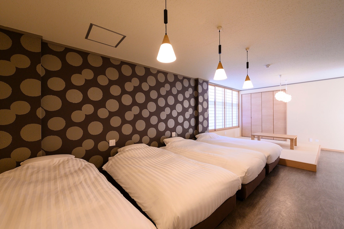 A spacious room features four beds dressed in white linens, arranged in a cozy layout. The walls are adorned with a patterned design, and soft lighting fixtures hang from the ceiling. A wooden table is positioned against the wall under a large window that allows natural light to enter.
