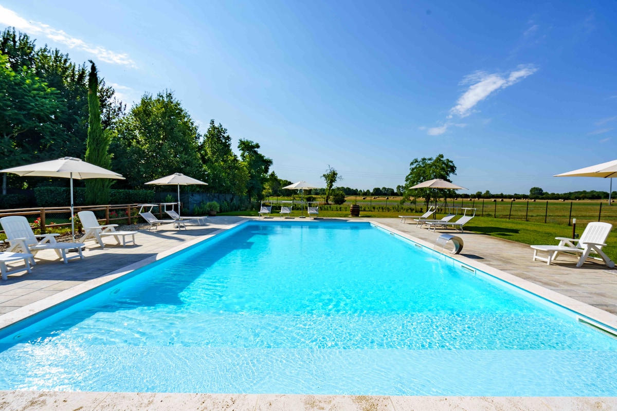 A spacious outdoor pool is surrounded by green gardens and shaded by umbrellas. Lounge chairs are placed around the pool for relaxation. The clear blue water reflects the sky, offering a serene space for swimming and leisure.