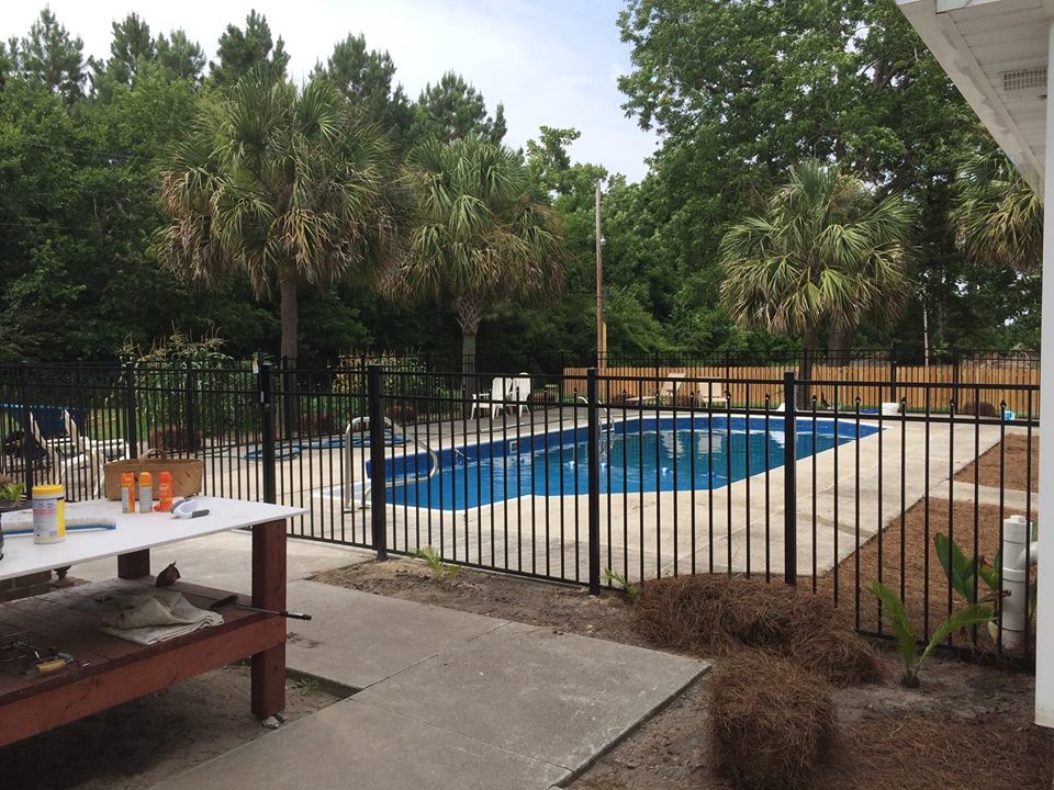 An in-ground swimming pool is surrounded by a black metal fence, with palm trees providing a tropical setting. The pool's clear blue water is visible, complemented by a sunlit patio area featuring lounge chairs. A concrete surface offers ample space for relaxation and leisure activities.