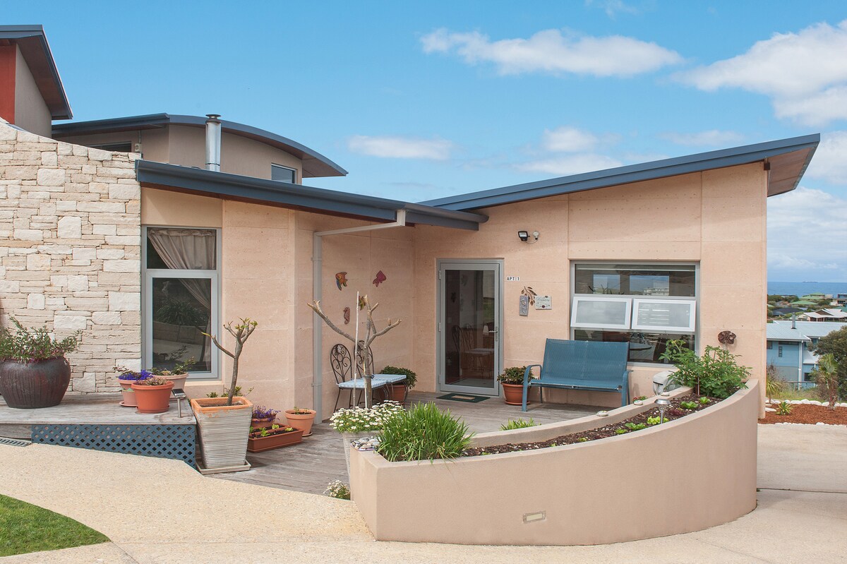 The exterior of the apartment features a unique rammed limestone design with a sloped roof. A small outdoor seating area is accented by potted plants. The entrance is adorned with a welcoming front door, and coastal views are visible in the background.